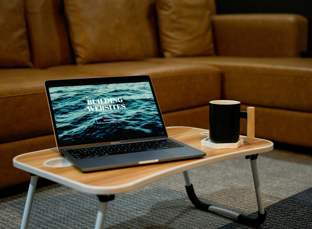 Laptop on a small table with “BUILDING WEBSITES” on the screen next to a coffee mug, symbolizing entrepreneurial focus and website development for business growth.